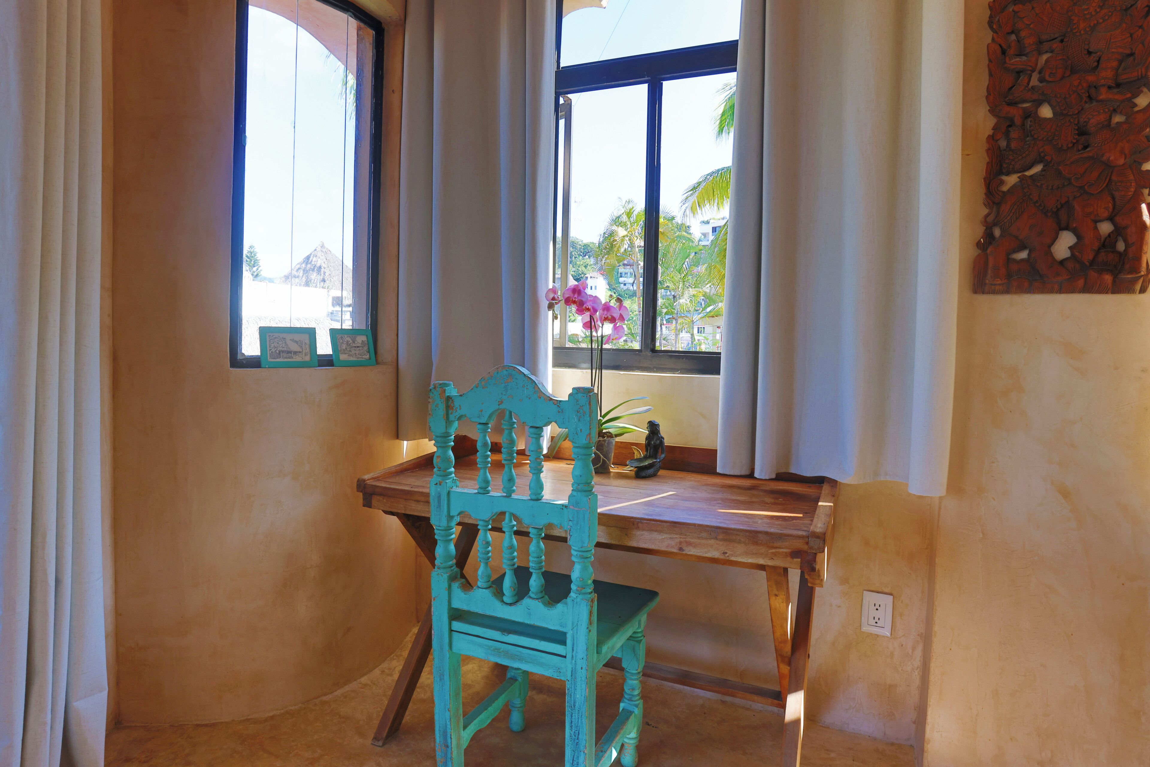 A room with a wooden desk, a turquoise chair, a window with curtains, and a wall-mounted decorative piece.
