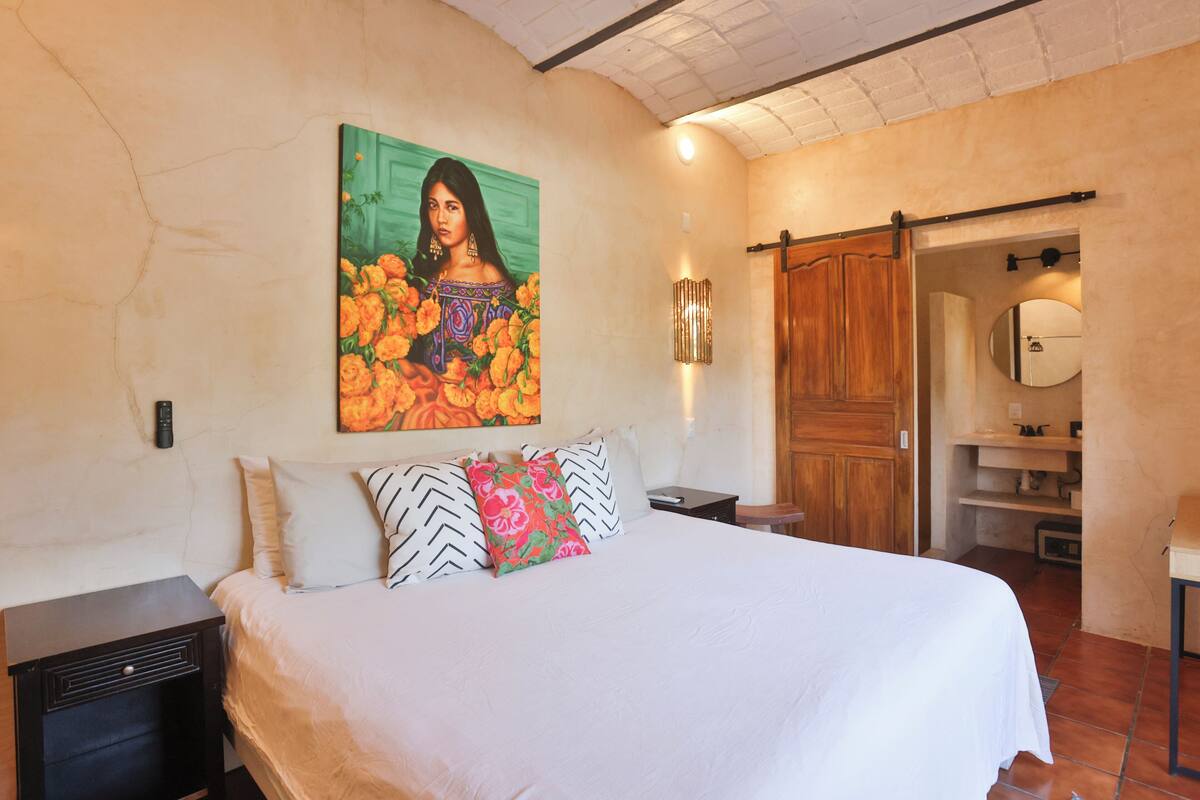 Un dormitorio con cama, almohadas, un cuadro colgado en la pared, una mesita de noche, una puerta de madera y un espejo.