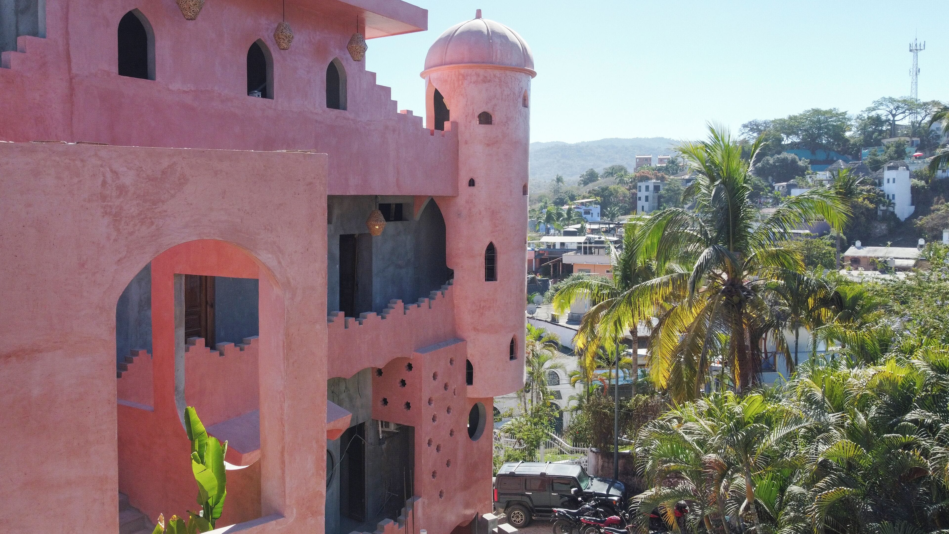 A pink building with arched windows and a tower, surrounded by palm trees and other greenery.