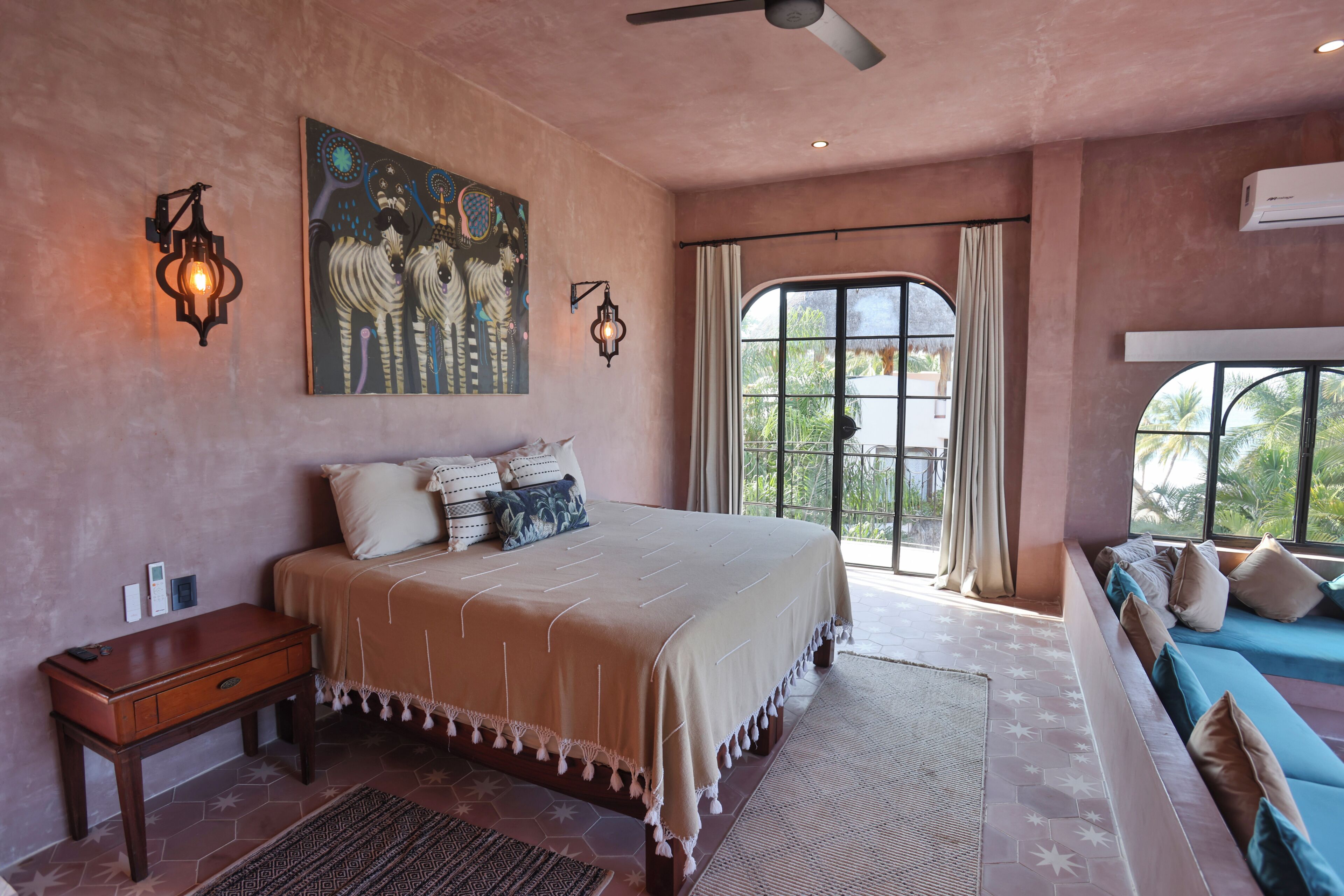 Quarto com cama grande, mesa de cabeceira, obra de arte na parede, ventilador de teto e janela com cortinas.