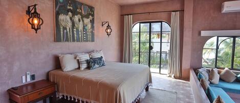 Quarto com cama grande, mesa de cabeceira, obra de arte na parede, ventilador de teto e janela com cortinas.