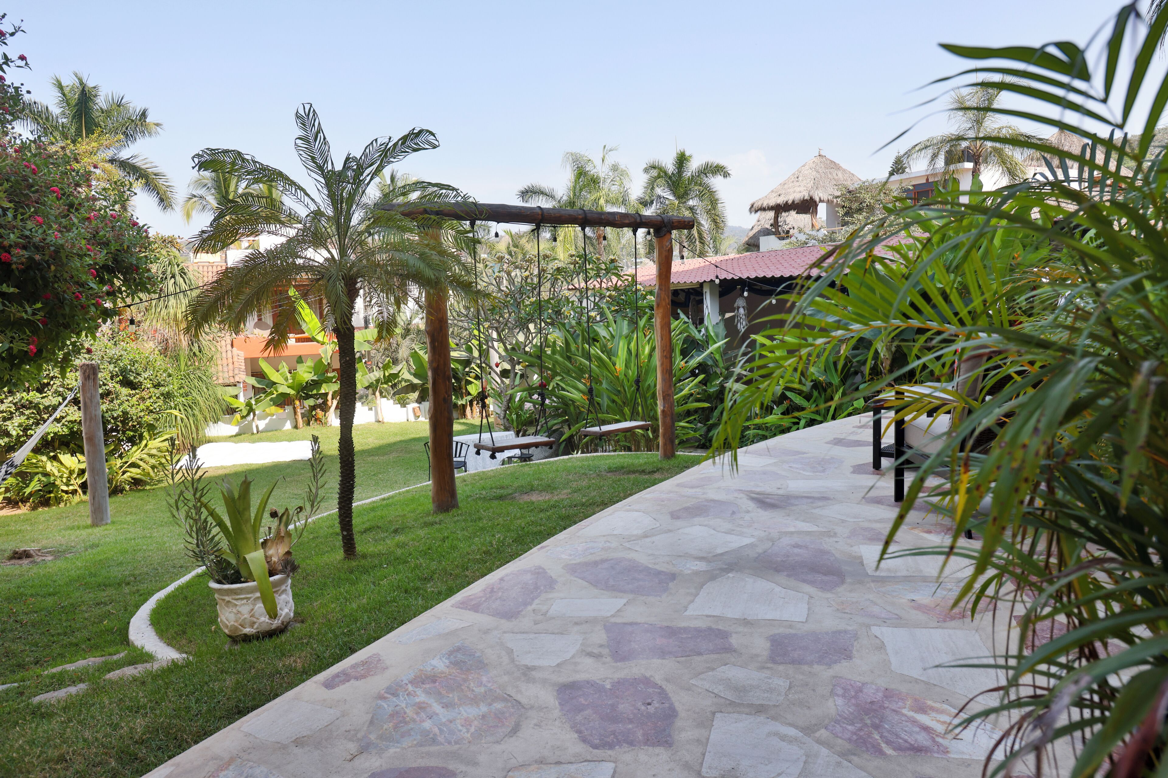 A stone pathway leading to a seating area with a thatched roof structure, surrounded by lush greenery and palm trees.
