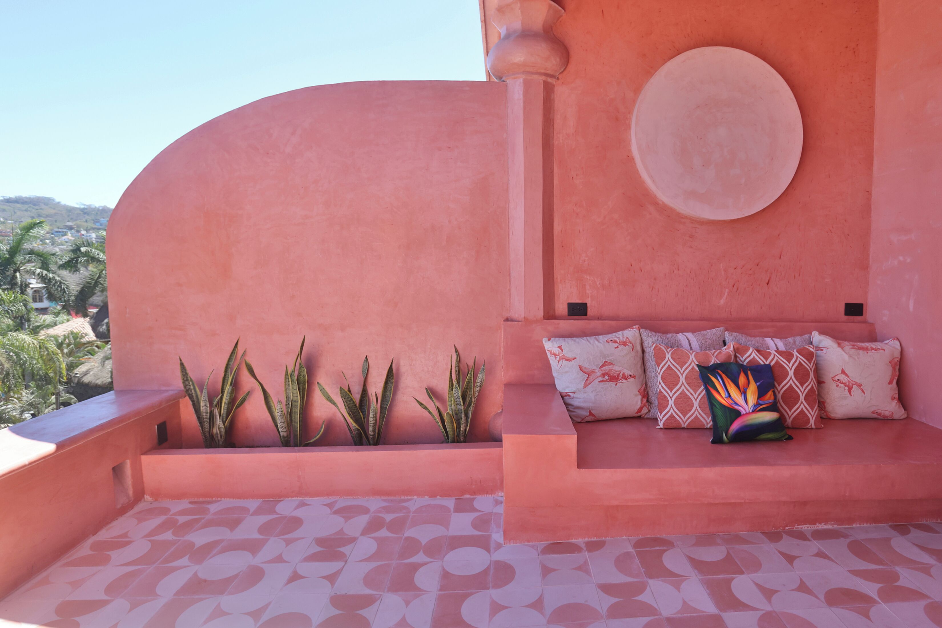 A pink-colored outdoor seating area with a sofa, cushions, and potted plants.