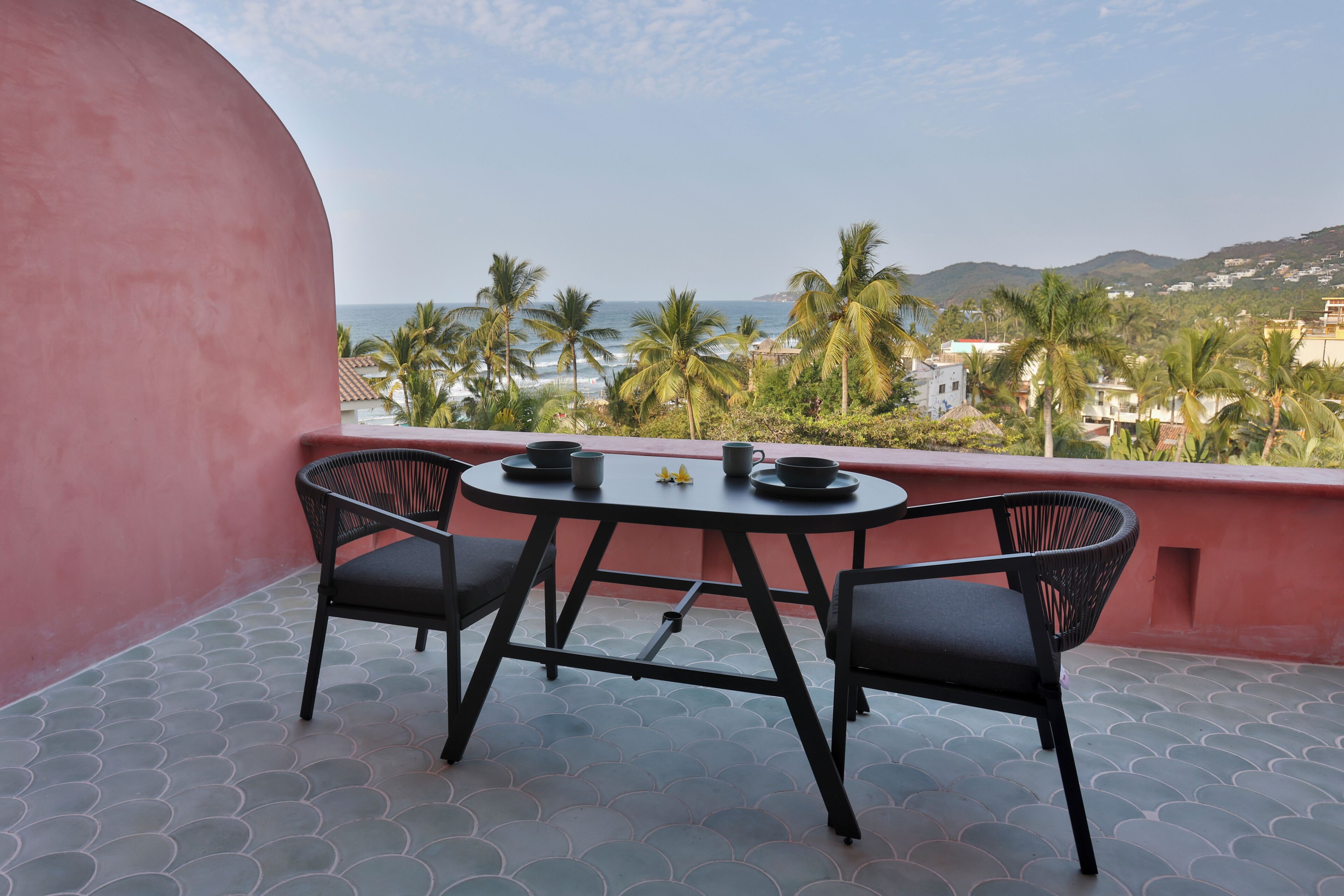 A balcony with a round table and two chairs, overlooking a coastal area with palm trees and buildings.