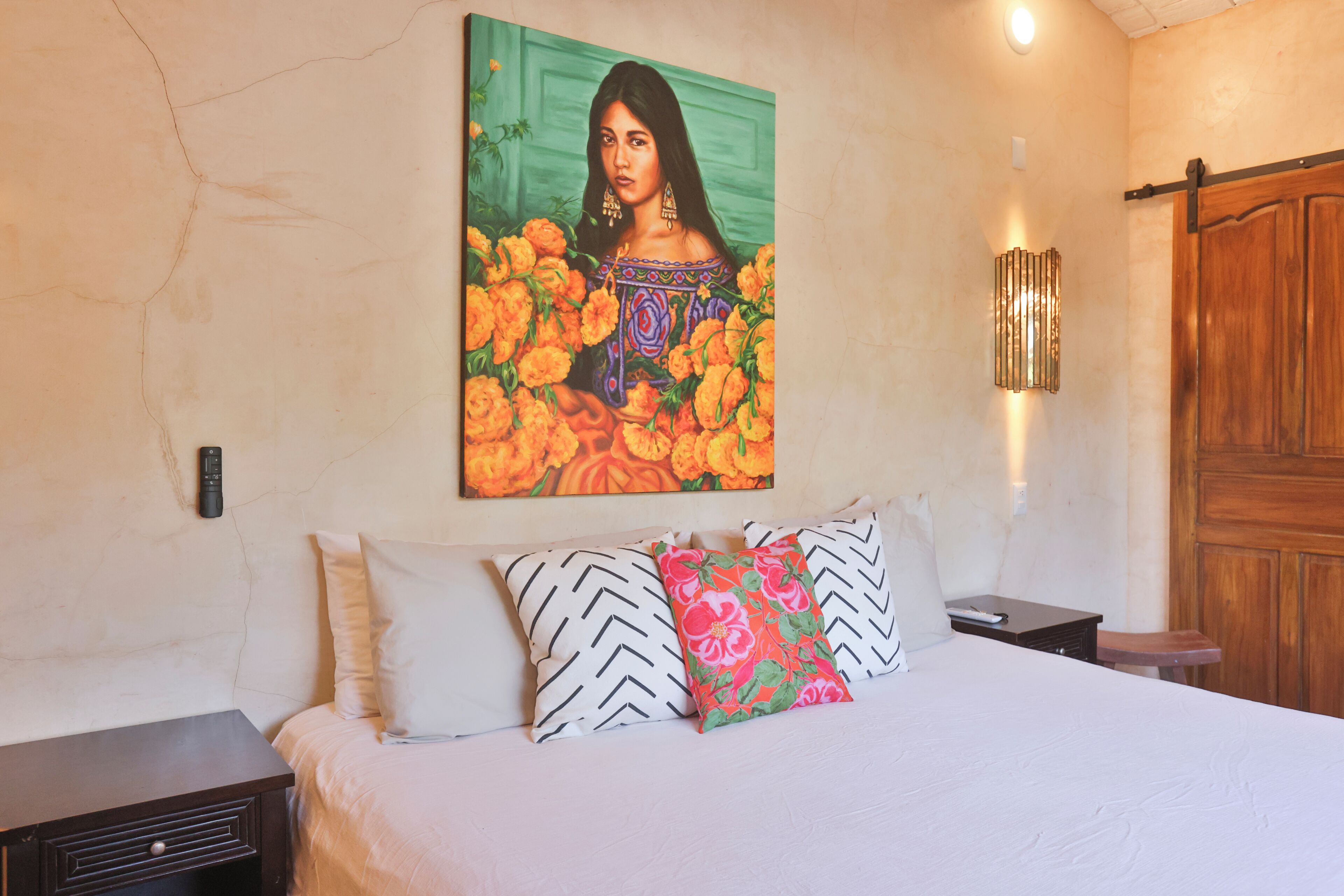 Una cama con almohadones estampados, un cuadro colgado en la pared, una mesita de noche y una puerta de madera.