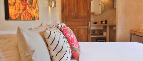 A bedroom with a bed, pillows, a wooden wardrobe, a mirror, and a bathroom visible in the background.