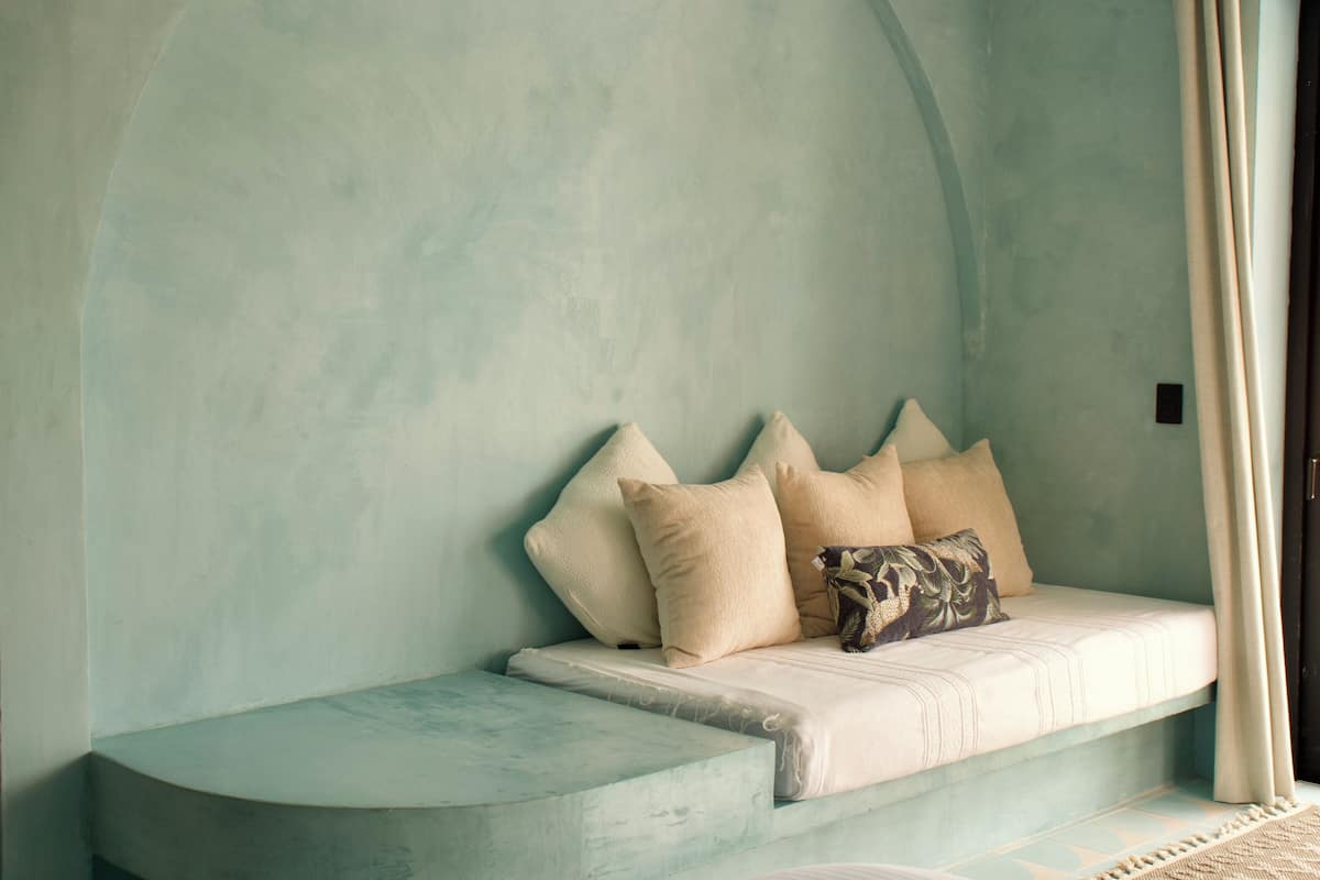 Una habitación con un banco curvo frente a una pared verde azulada, una ventana con cortinas y un cojín estampado.