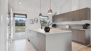 Una cocina moderna con armarios grises, una isla central, iluminación colgante y vistas a través de un ventanal.