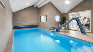 Una piscina interior con un tobogán azul, una escalera y vistas a un comedor a través de un cristal.