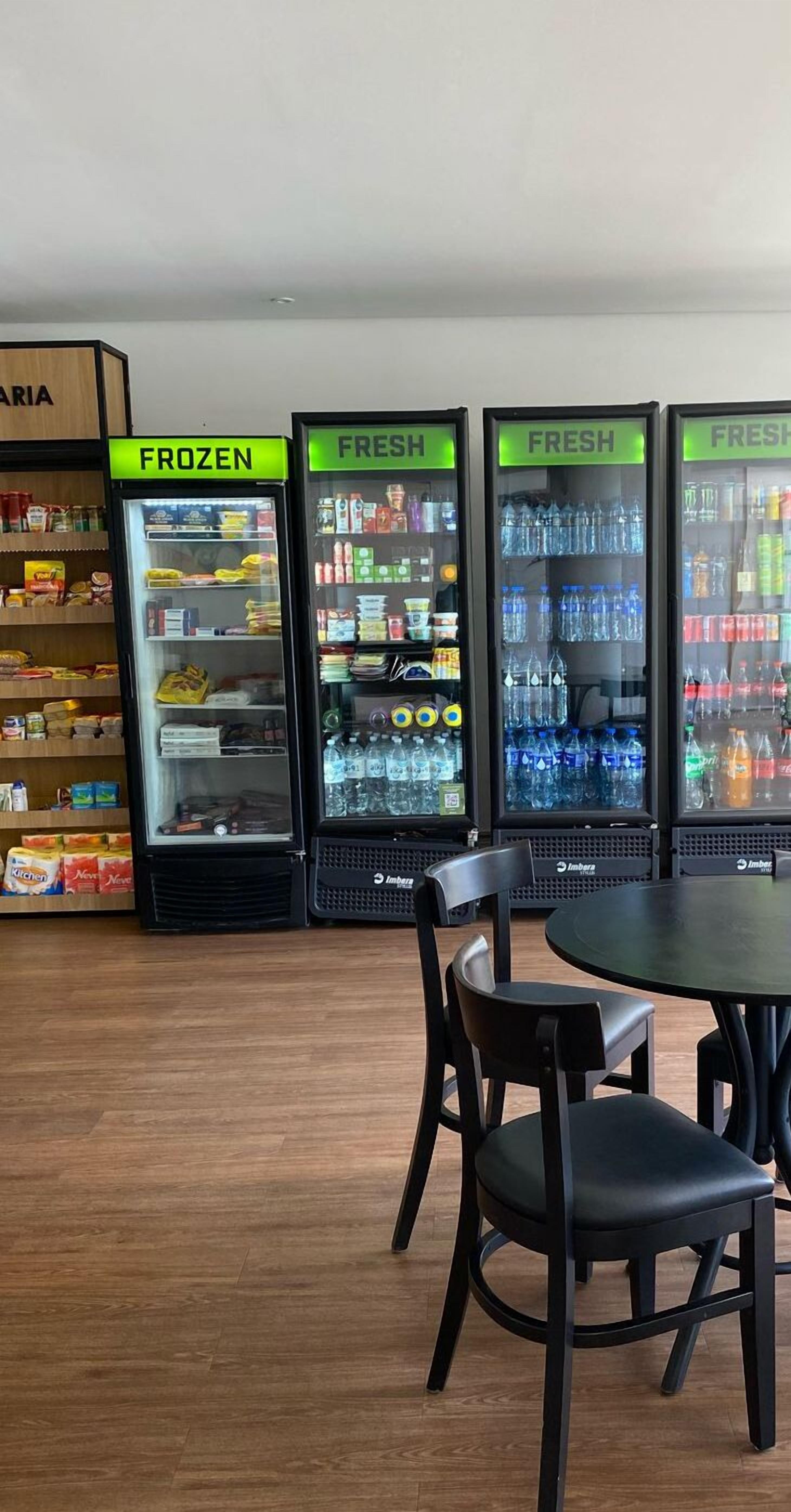 A row of beverage dispensers labeled 'FROZEN' and 'FRESH' in a food service area with wooden flooring and black chairs.