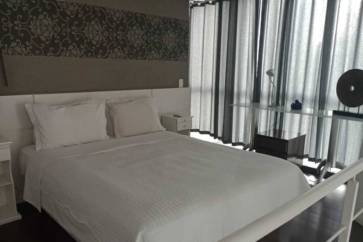 Ein ordentlich bezogenes Bett mit weißen Leinen in einem modernen Zimmer, das ein gemustertes Kopfteil, einen Nachttisch und ein Fenster mit durchscheinenden Vorhängen hat.