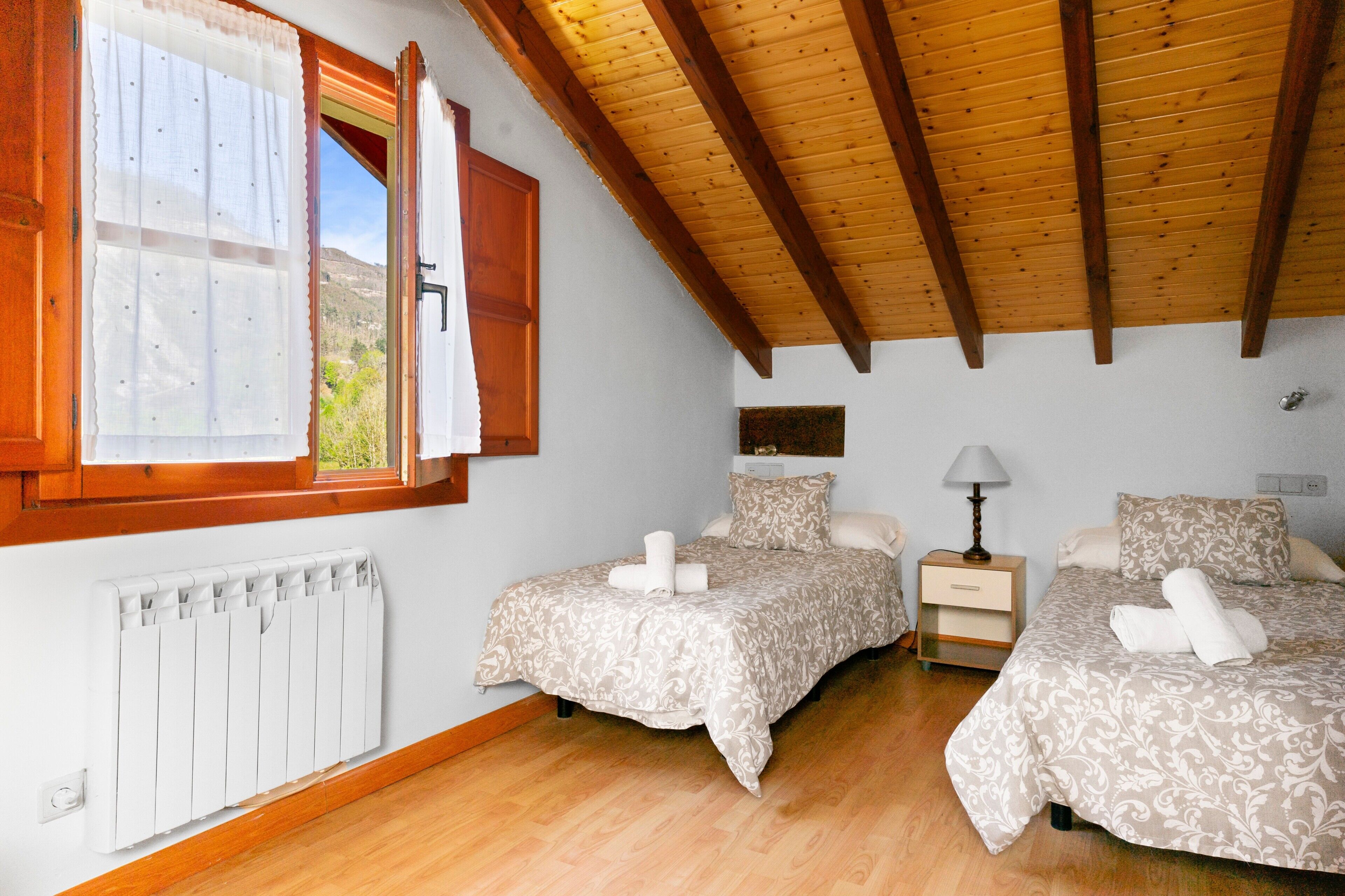 Une chambre avec deux lits, une fenêtre avec des rideaux, un plafond en bois, une table de chevet avec une lampe et un radiateur.