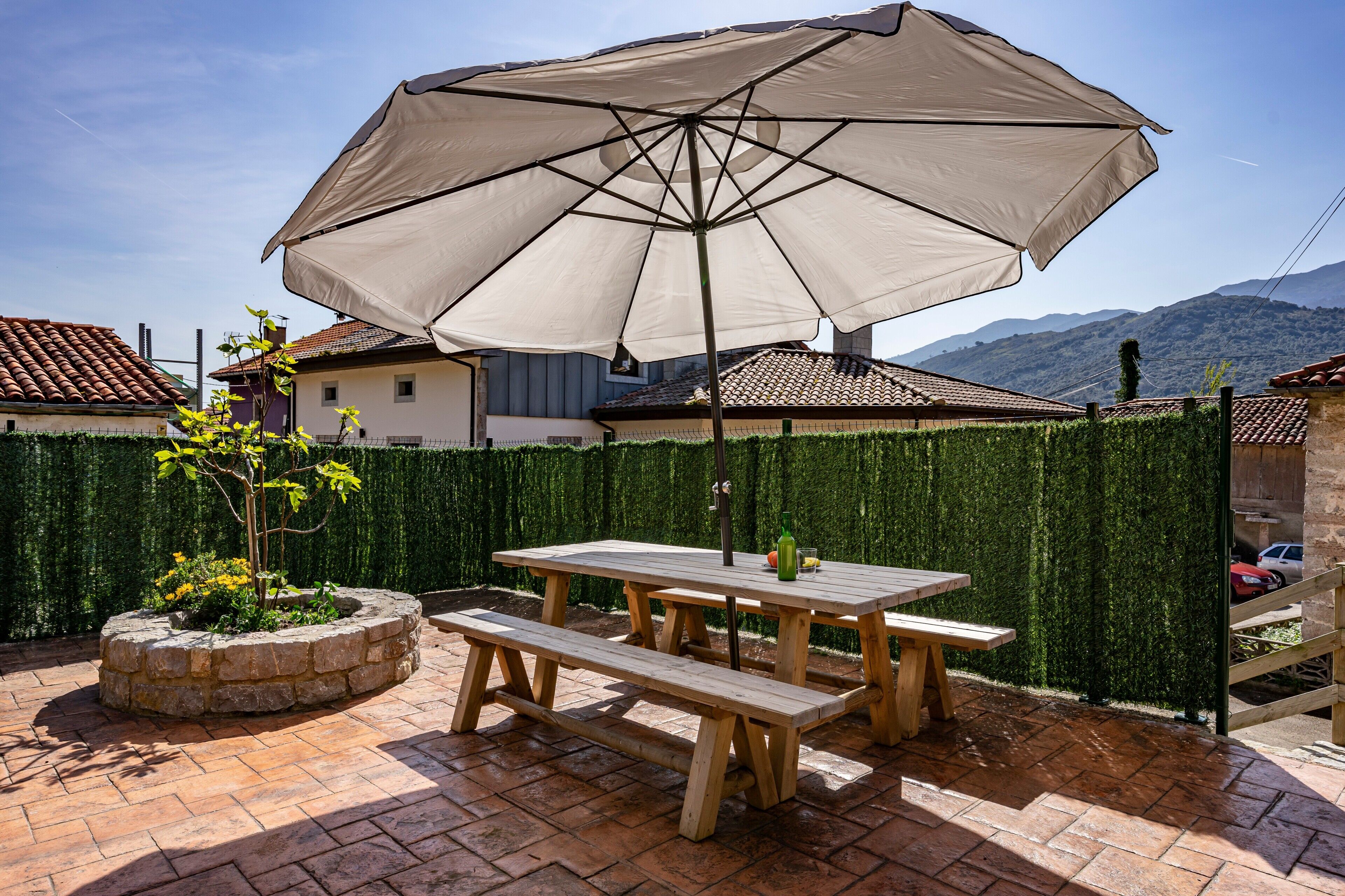 An outdoor patio with a wooden table and benches, a large umbrella, and a stone planter with a tree.
