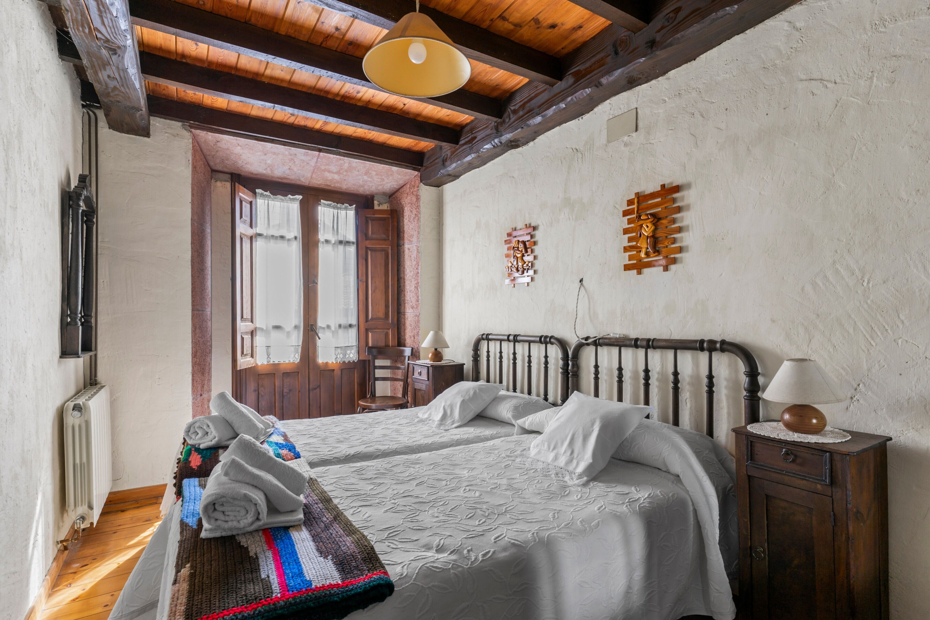 Un dormitorio con dos camas, vigas de madera, un radiador, una mesita de noche de madera con una lámpara y una ventana con cortinas.