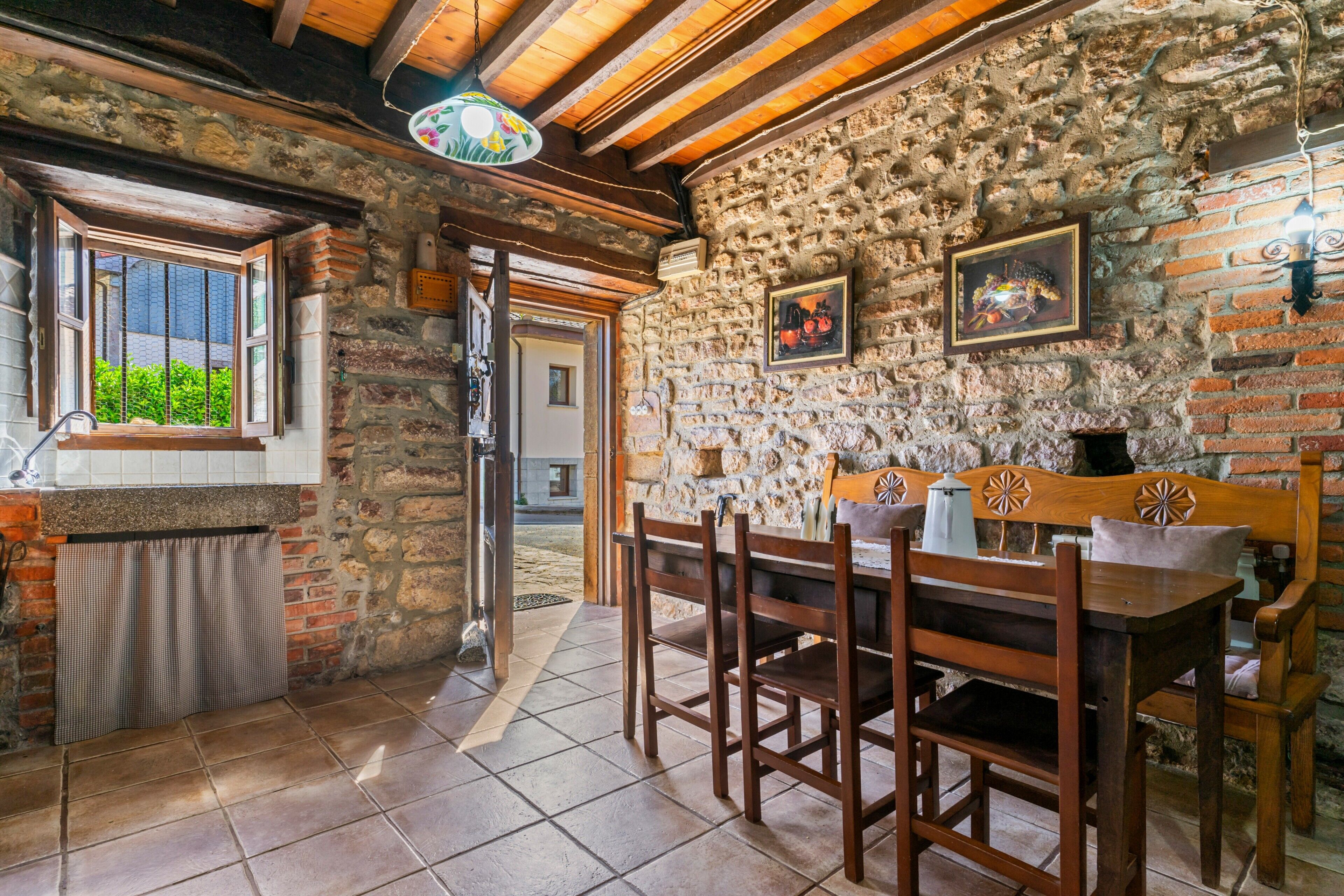 Un interior rústico de piedra con una mesa y sillas de madera, una ventana con vistas, y una puerta que da al exterior.