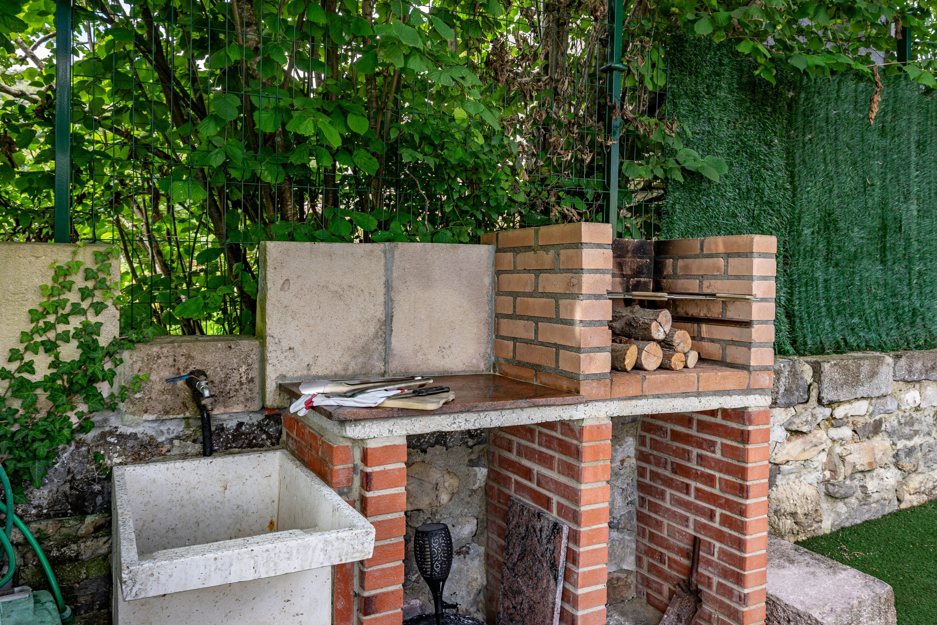 Un four en briques installé à l’extérieur, avec un mur en pierre, un évier en béton et un tuyau d’arrosage.