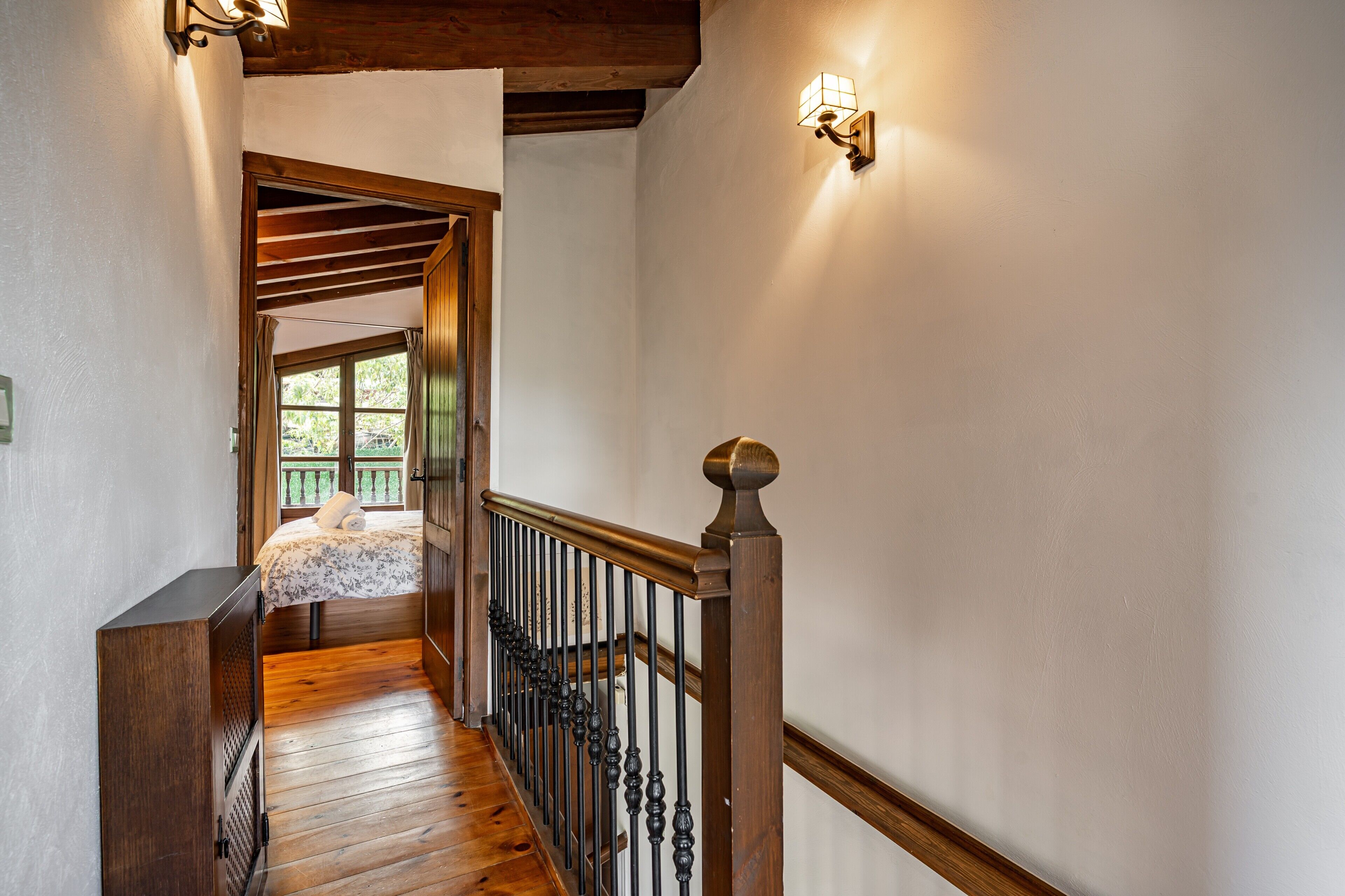 A wooden staircase with a railing, a bed with a floral bedspread, and a window with a view of greenery.