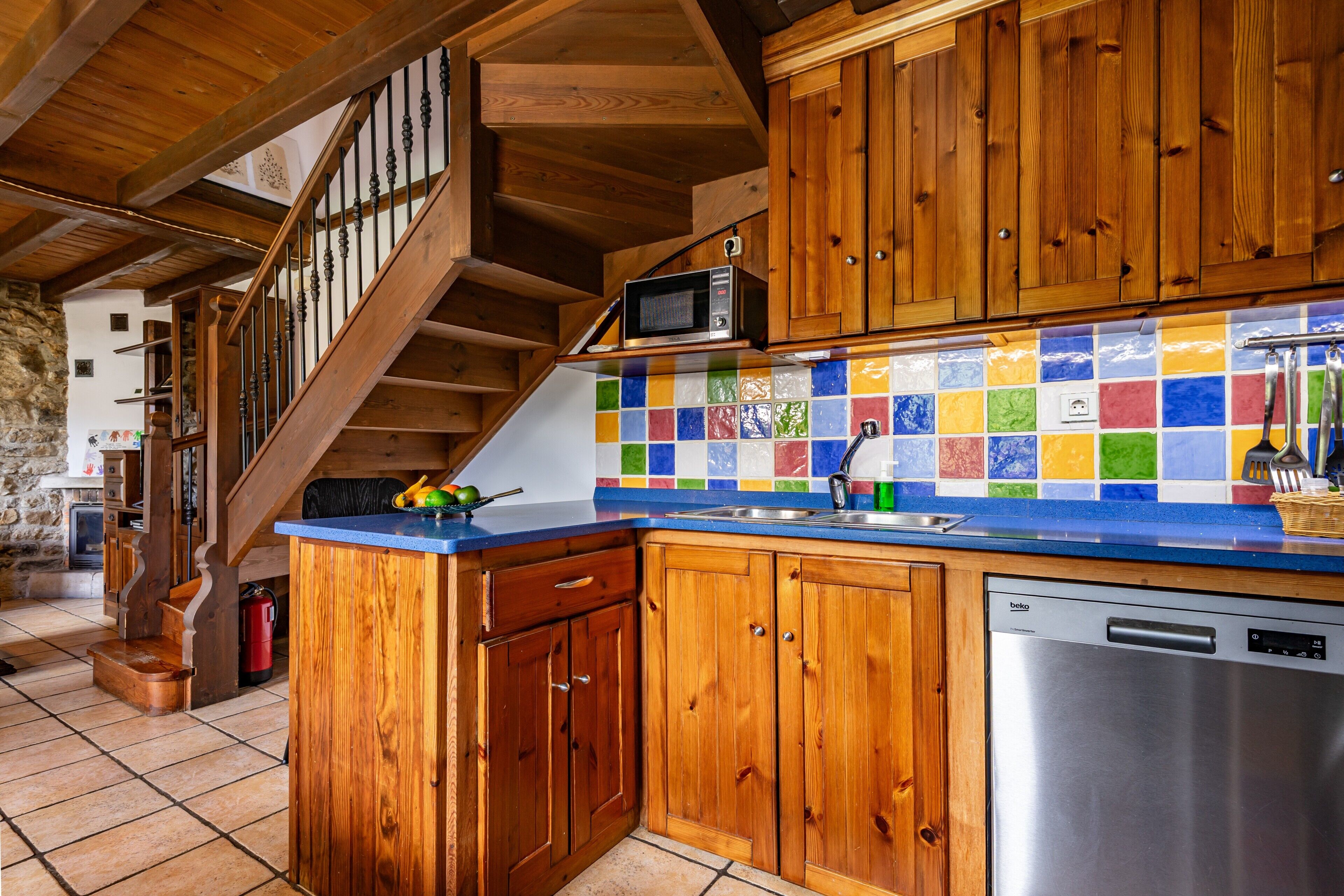 Eine Küche mit Holzschränken, einem Edelstahl-Geschirrspüler, einer farbenfrohen Fliesenrückwand und einer Holztreppe.