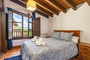 Une chambre avec un plafond en bois, un grand lit recouvert d’une couvre-lit à motifs floraux, une table de chevet en bois et une fenêtre donnant sur un immeuble et des arbres.