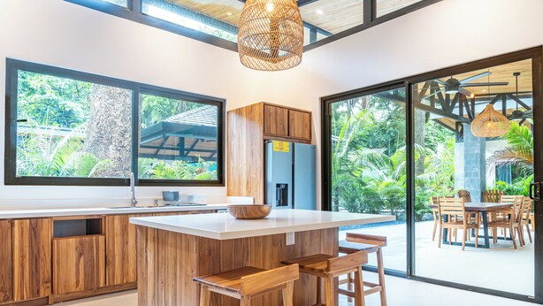 A modern kitchen with wooden cabinetry, a large island with bar stools, and large windows offering a view of lush greenery.