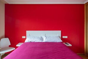 Una habitación con una cama, mesitas de noche blancas, una silla blanca y una pared roja.