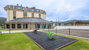 A playground with a frog-shaped swing set and a seesaw, a building with multiple windows and a sloped roof, and a fenced area with a bench. - Apartment 'San Miguel Reinante' with Mountain Views, Pool and Wi-Fi (SAN MIGUEL DE REINANTE)