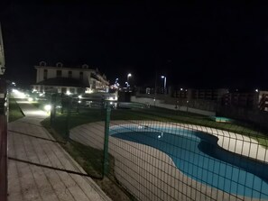 Vista nocturna de una zona de piscina con valla, un camino y edificios al fondo.