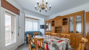 Comedor con una mesa dispuesta para cuatro personas, sillas de madera con cojines verdes, una araña, un aparador grande con puertas de vidrio y un televisor sobre un mueble.