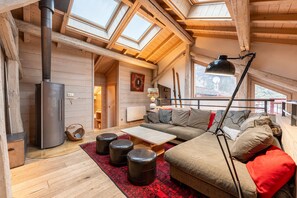 A cozy loft-style room with a fireplace, wooden beams, a sofa, ottomans, and a large window.