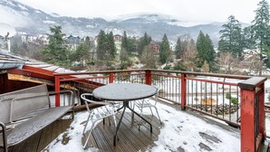 Una terraza de madera con sillas y una mesa de metal, con vista a un paisaje nevado con casas y árboles.