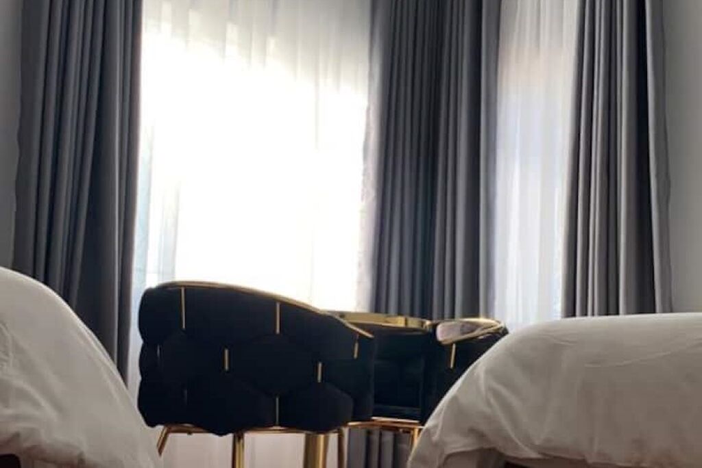 Hotelkamer met twee bedden, een donkerbeklede fauteuil en uitzicht op de stad door grote ramen met dunne gordijnen.