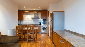 Cocina compacta con armarios de madera, horno empotrado, lavadora y comedor con sillas.