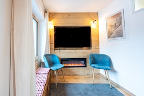 Een gezellige kamer met een open haard, twee blauwe stoelen, een geruit tafeltje en een grote televisie bevestigd aan een houten wand.