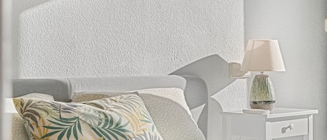 A bedroom with a textured wall, a bed with patterned pillows, a bedside table with a lamp, and a nightstand with a vase.