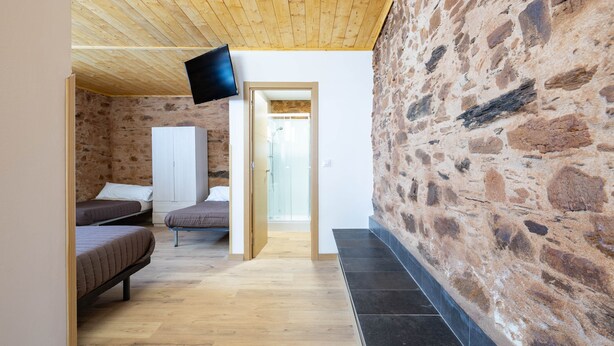 A room with a stone wall, wooden flooring, a bed, a wardrobe, and a flat-screen TV mounted on the wall. - 'Baloco' Apartment with Shared Pool, Terrace, and Wi-Fi (Monforte de Lemos)