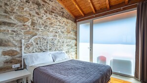 Une pièce avec un mur en pierre, un lit doté d’une tête de lit en métal blanc, un banc et une vue sur l’extérieur, visible à travers une porte vitrée.