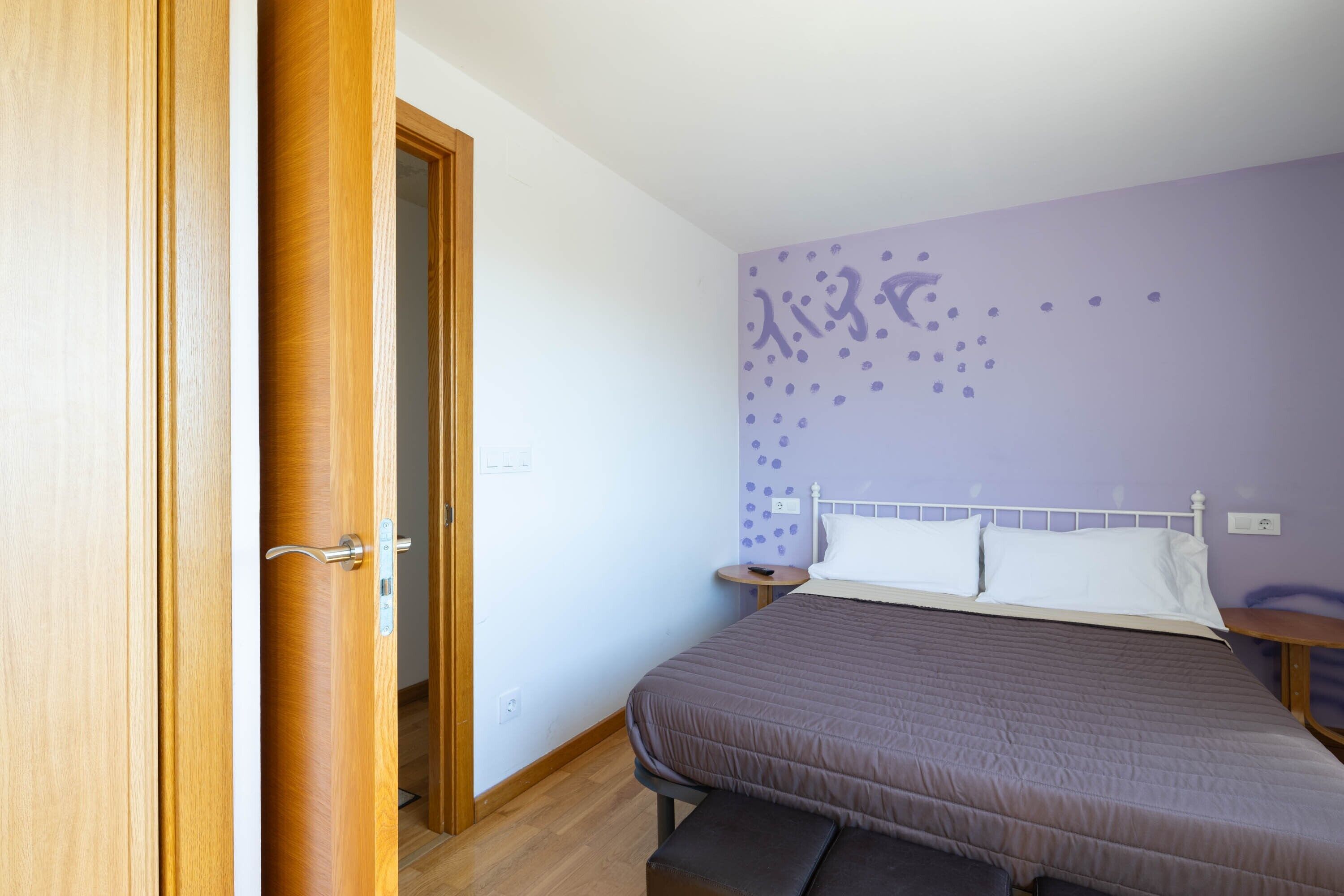 A bedroom with a bed, a wooden door, a nightstand, and a wall with a purple pattern.