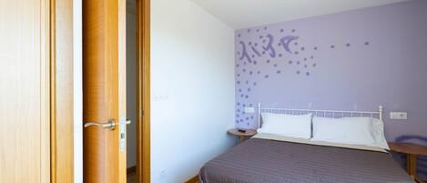 A bedroom with a bed, a wooden door, a nightstand, and a wall with a purple pattern.