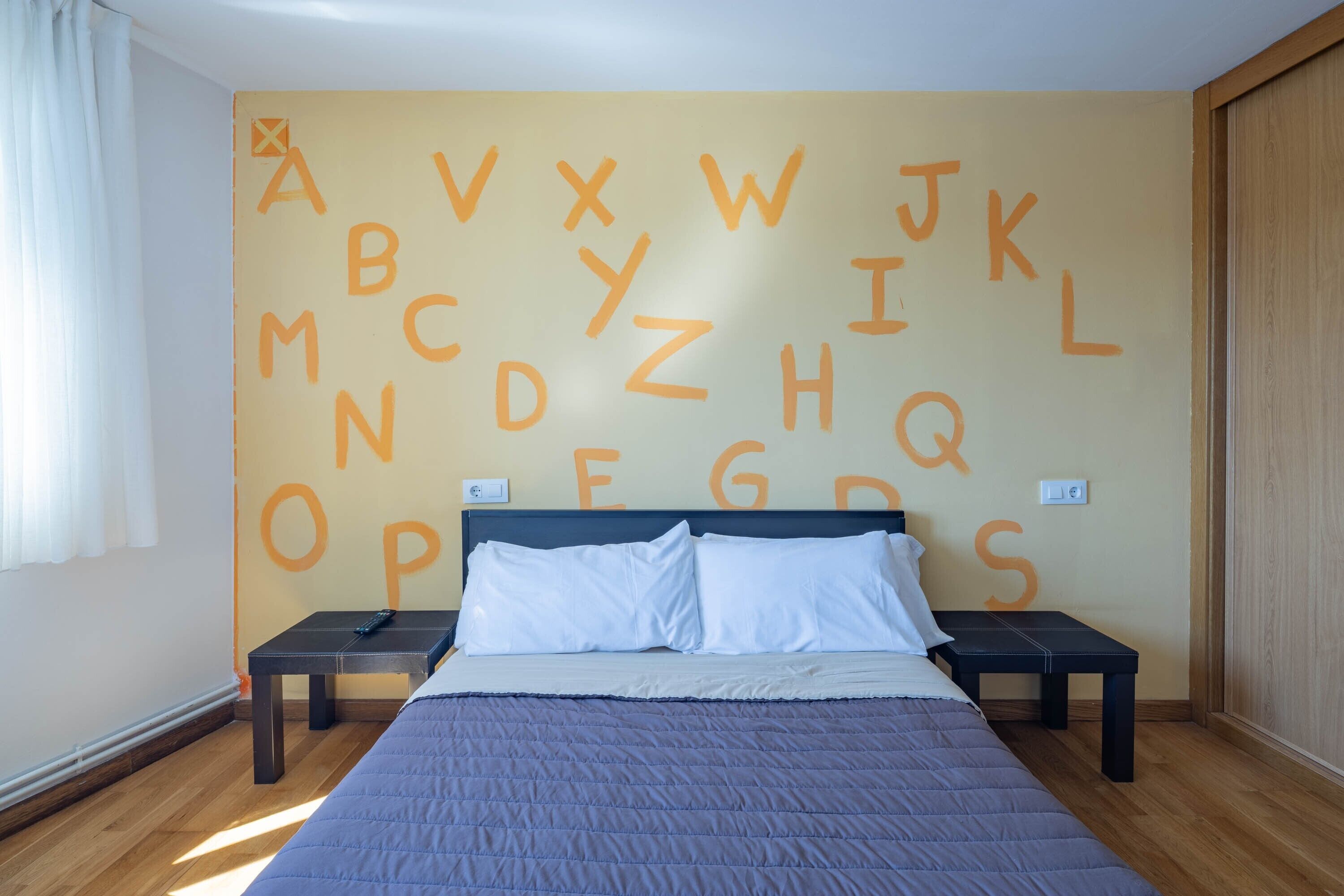 A bedroom with a bed, two bedside tables, and a wall with large alphabet letters.