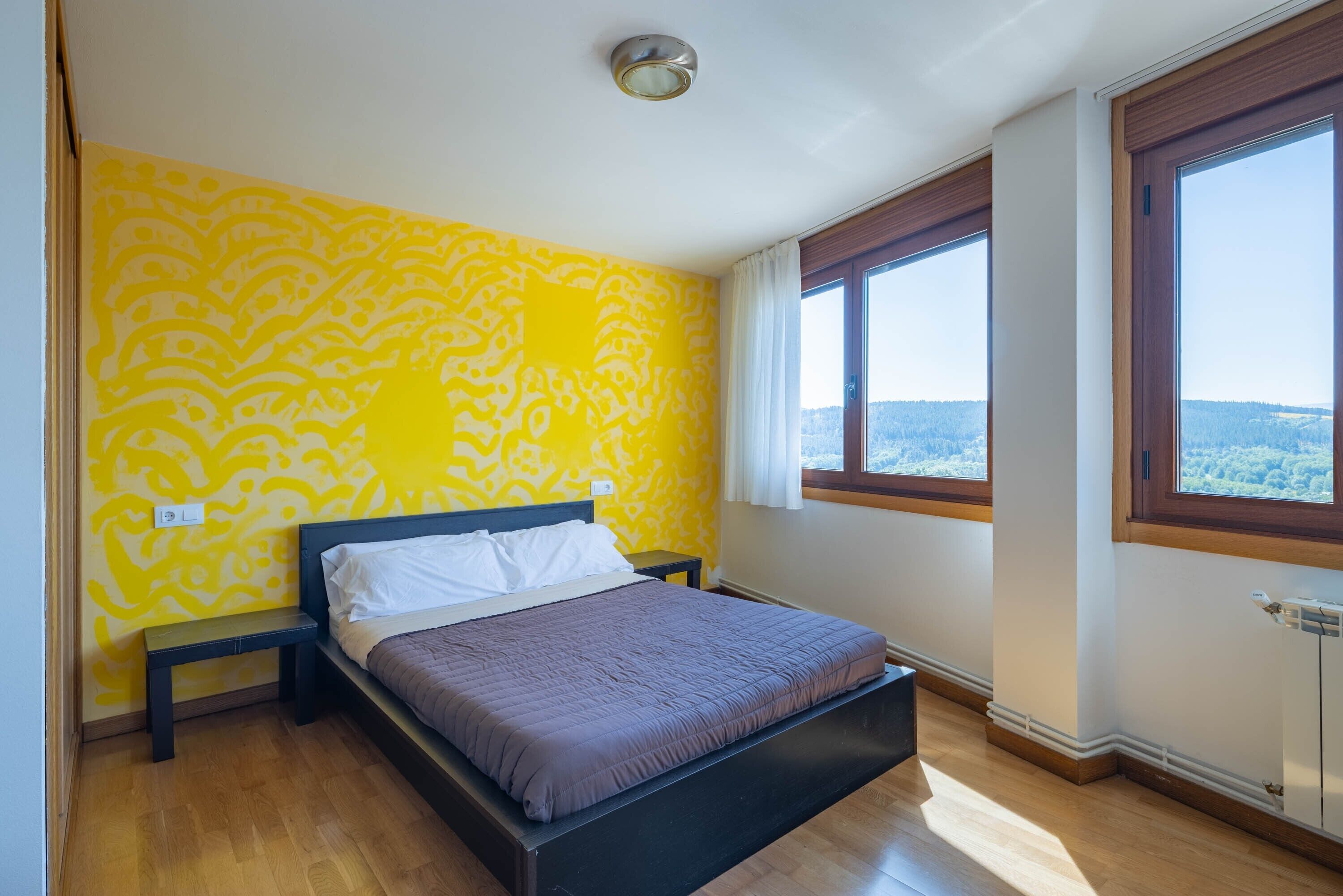 A bedroom with a bed, a nightstand, a window with a view, and a patterned wall.