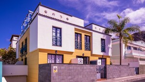 Un edificio de dos pisos con fachada amarilla y blanca, un acceso con reja y un balcón visible.