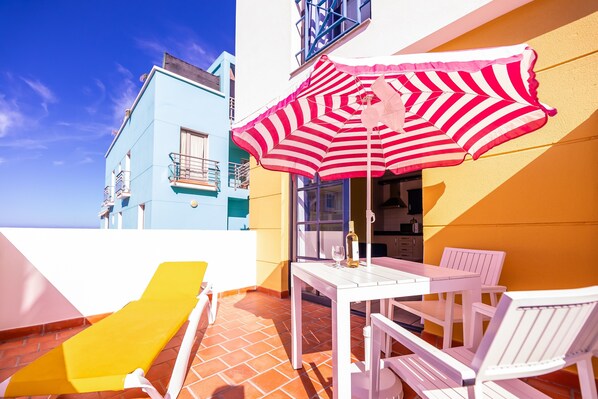 A terrace with a white table and chairs, a striped umbrella, and a yellow lounge chair.