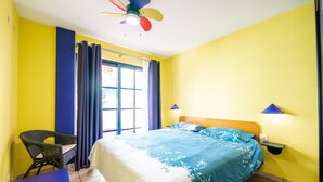 Una habitación con cama, una silla, una mesa, ventilador de techo y ventana con cortinas.