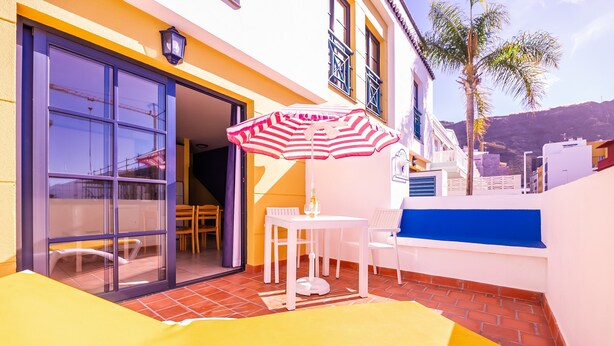 Un patio soleado con una silla reclinable amarilla, un parasol a rayas y una mesa con sillas.