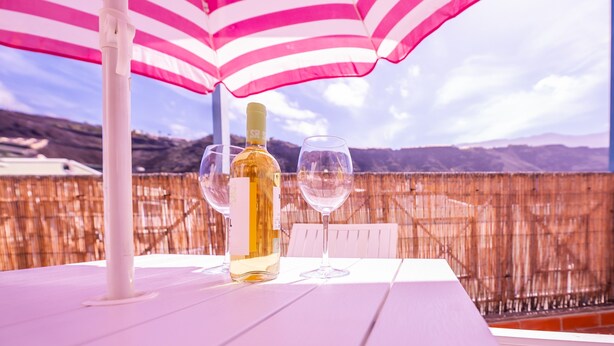 Una mesa con una botella y dos vasos bajo un paraguas a rayas, con vistas a montañas al fondo.