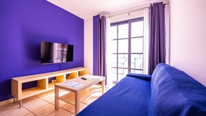 Una habitación con un sofá azul, un televisor instalado en la pared, una mesa de madera y una ventana con cortinas moradas.