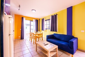 A living room with a blue sofa, a wooden dining table with chairs, and a kitchen area with a refrigerator.