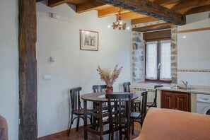 A dining area with a round table, chairs, a vase with dried plants, a chandelier, and a kitchen with a stone wall and window. - Country House 'Lope' with Mountain View, Private Garden and Wi-Fi (Lores)