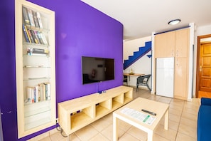 Una sala de estar con televisión, estanterías, un banco de madera, una mesa con control remoto y una escalera.