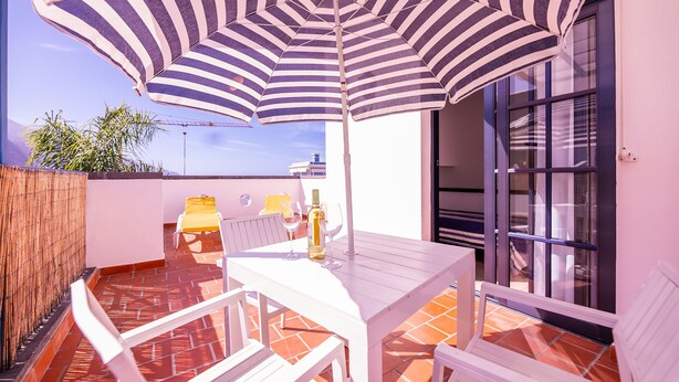 Un patio con un paraguas a rayas, muebles blancos y suelo de baldosas.
